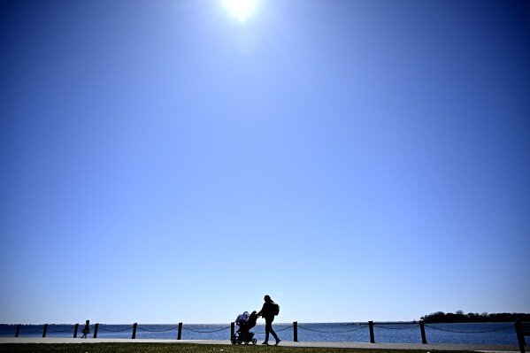 A blue sky shines below the sun as a person walks with an infant along the shores of Lake Ontario, a day before a total solar eclipse will be visible in Kingston, Ontario, Sunday, April 7, 2024. (Justin Tang/The Canadian Press via AP)